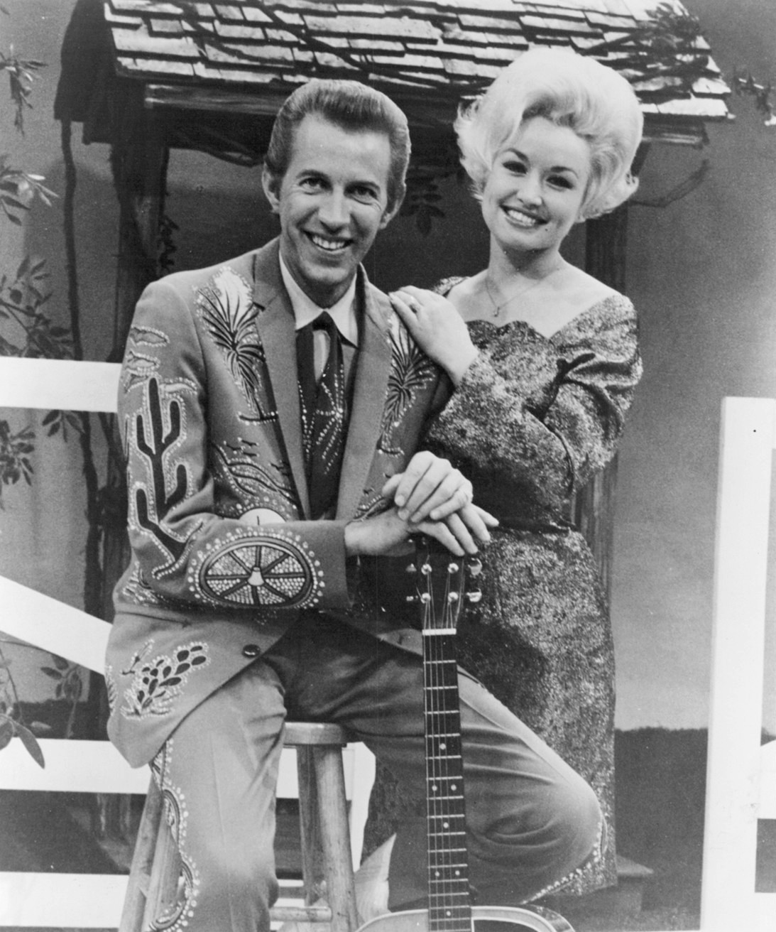 Black-and-white 1969 press photograph: Dolly Parton standing beside a seated Porter Wagoner, who holds a guitar on a rustic studio set.